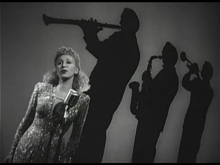 Canted angle shot. Singer in foreground and silhouettes of horn players in background. Image appears tilted.